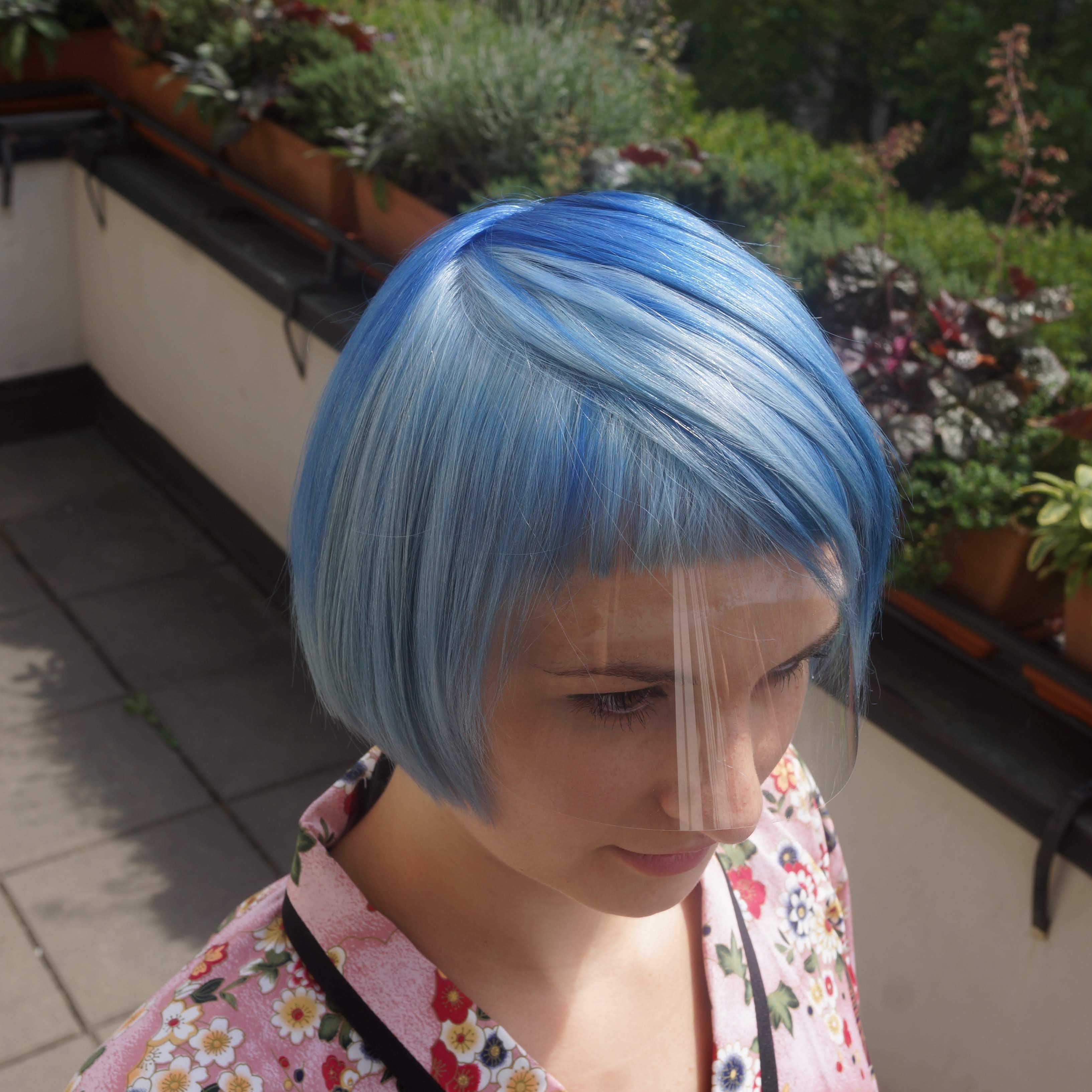 Person with blue hair wearing a face shield on a patio with plants and a bench.