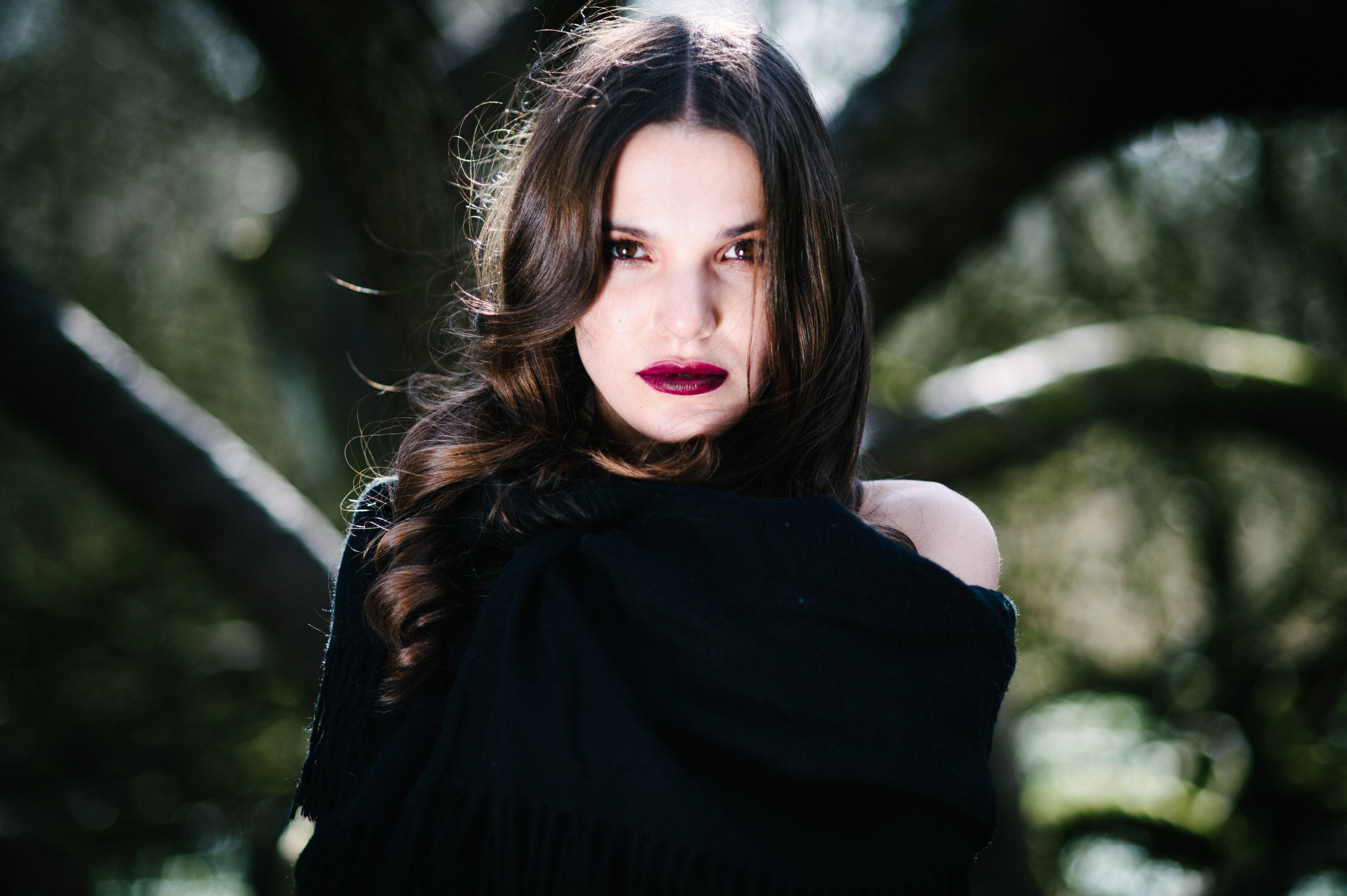 Woman with dark hair and red lipstick standing against a blurred natural background