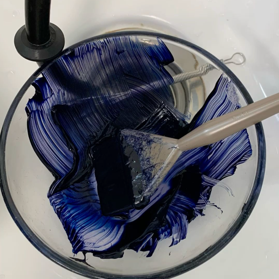 Blue paint being stirred in a glass bowl with a metal spatula.