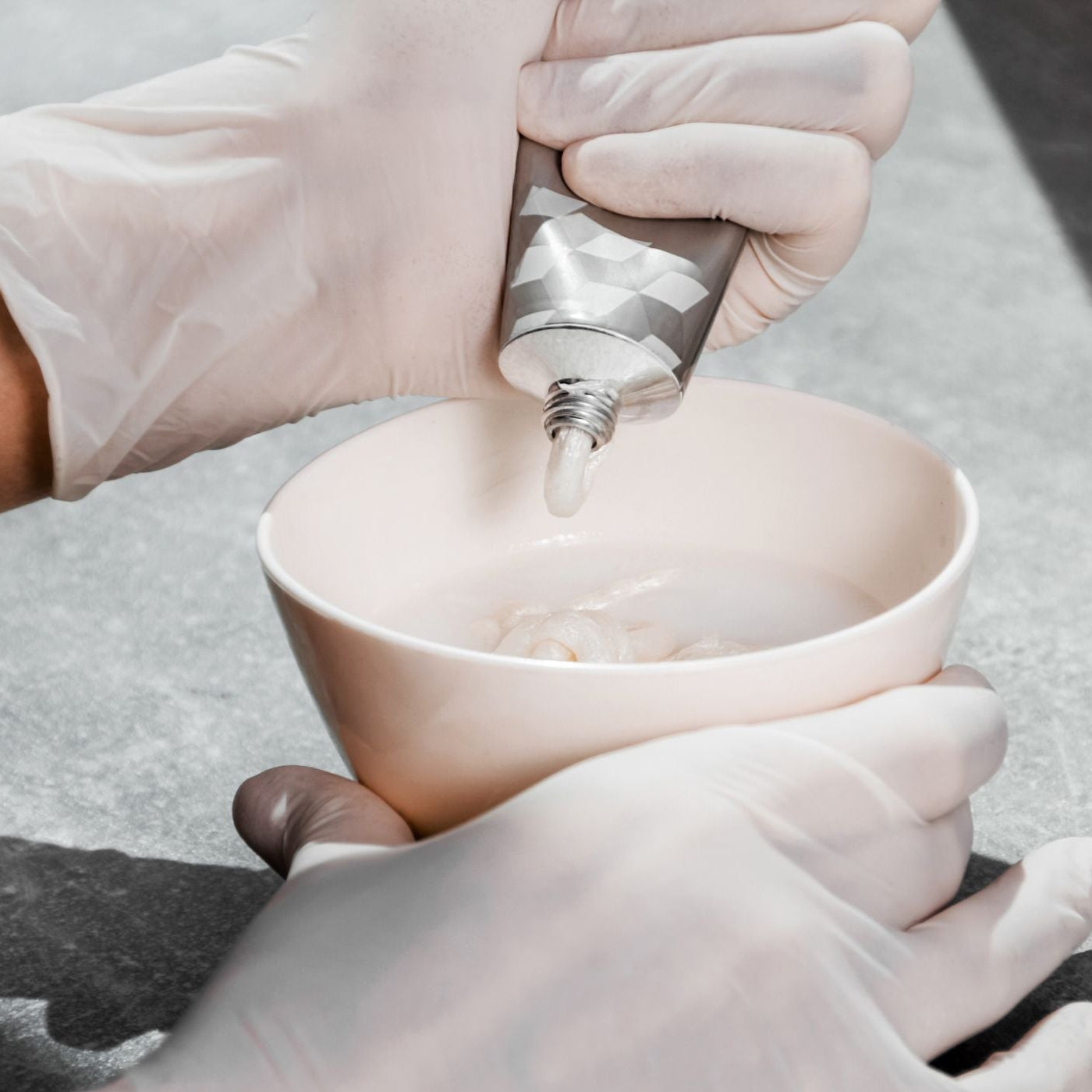 Person wearing gloves pouring hair dye cream from a tube into a pink bowl.