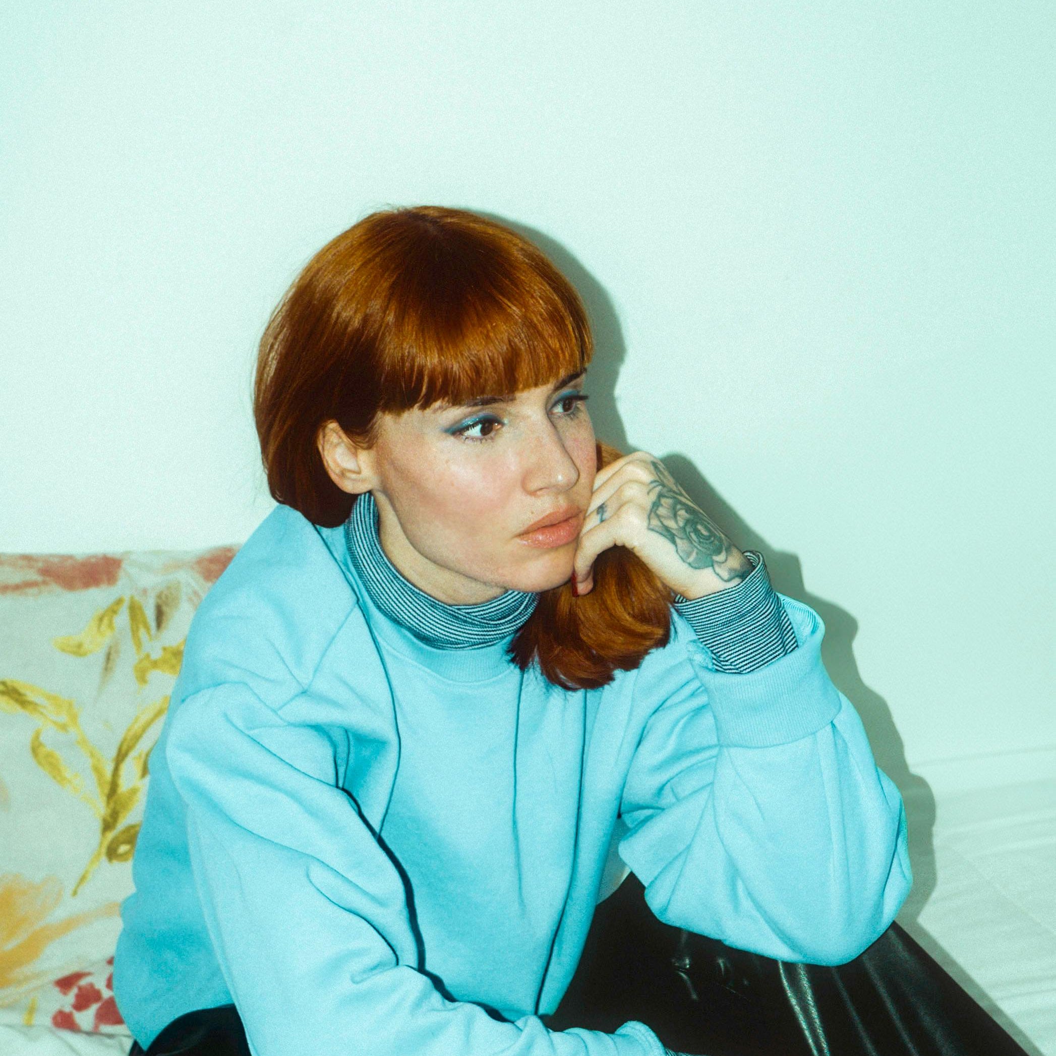 Person wearing a light blue jacket sitting on a bed with a white wall in the background